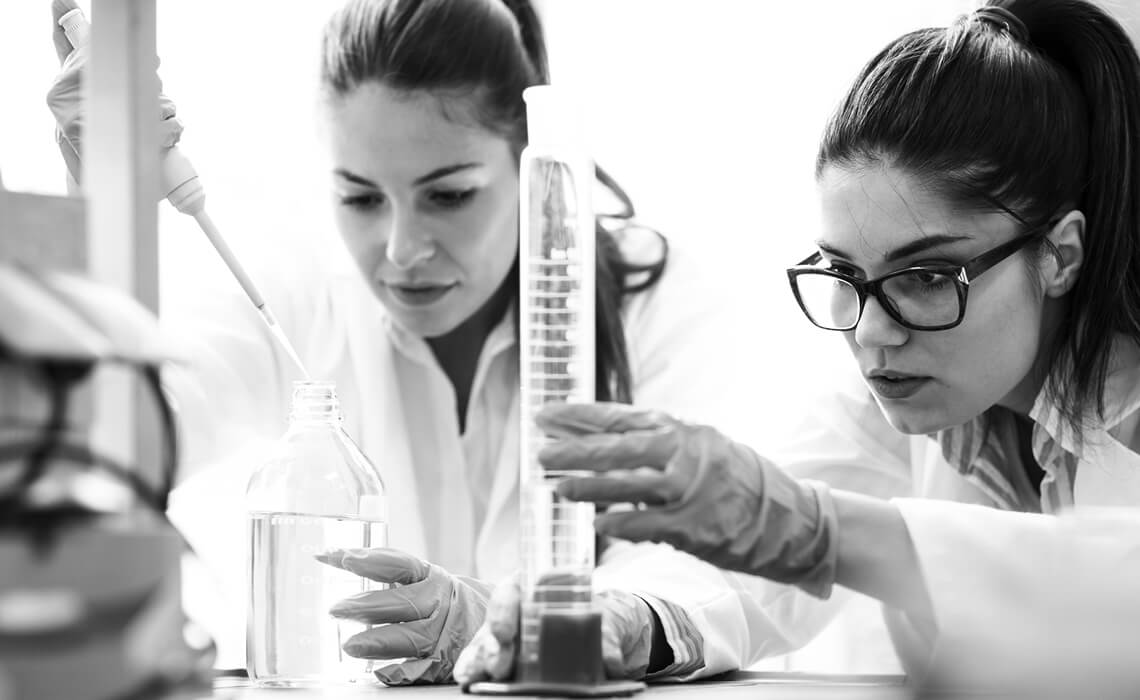 Two women in lab coats - one using a dropper to add liquid to a bottle and the other measuring liquid in a beaker